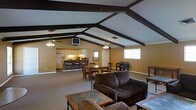 This single-story space makes a striking first impression with its soaring vaulted ceilings adorned with bold, dark-stained exposed wood beams that run the length of the roofline, lending the interior a rustic yet airy character. The open-concept layout seamlessly connects the living, dining, and kitchen areas into one expansive, flowing space.

The living area is anchored by plush, dark brown upholstered sofas arranged around a sturdy wood-and-stone coffee table, creating a comfortable and welcoming gathering space. Ceiling fans are thoughtfully positioned throughout to ensure consistent airflow across the generous open floor plan.

The dining area features a classic wooden dining table with matching chairs, positioned centrally between the living space and the kitchen. The kitchen is well-appointed with white cabinetry, a full-size refrigerator, and a raised breakfast bar with barstool seating, making it both functional and social.

Natural light filters in through multiple windows, brightening the warm golden-toned walls and neutral carpeted floors. A wall-mounted television is integrated into the space near the kitchen, adding to the home's casual, lived-in comfort. The cohesive design and open sightlines throughout create a spacious, practical, and welcoming environment ideal for everyday living and entertaining.