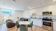 This contemporary high-rise residence offers a thoughtfully designed, open-concept layout spread across a single floor, with light wood flooring and clean white walls throughout creating a bright, cohesive aesthetic. The kitchen is well-appointed with white shaker cabinetry, granite countertops, stainless steel appliances including a full-size refrigerator, range, built-in microwave, and dishwasher — all arranged in an efficient layout that flows seamlessly into the dining and living areas. A round marble-top dining table anchors the combined dining space, while the living area features floor-to-ceiling windows that flood the room with natural light and frame sweeping views of the Detroit skyline. The bedroom is a serene retreat, also benefiting from large windows with expansive city views, white walls, and light hardwood flooring. A generously sized walk-in closet with wire shelving provides ample storage. The entry corridor leads past a vanity with dark wood cabinetry and stone countertop, hinting at a well-appointed bathroom nearby. Recessed lighting throughout enhances the modern, polished feel of the space. The combination of contemporary finishes and dramatic urban views makes this a compelling city residence.