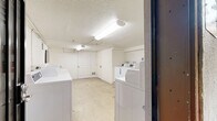 Located at 3902–3920 Lenawee Ave in Culver City, California, this shared laundry facility serves the surrounding residential community. The single-floor utility room is efficiently laid out to accommodate multiple top-loading washing machines and dryers, providing ample capacity for residents. The interior features textured white ceilings with overhead fluorescent lighting, white-painted walls, and a concrete floor, reflecting a practical, no-frills design focused on function. A standard entry door provides easy access to the space. Safety equipment, including a fire extinguisher, is visibly present. The facility is set within a well-maintained mid-century style residential complex that features a welcoming central courtyard with tall palm trees, decorative drought-tolerant landscaping with red mulch and river rock, and wide concrete walkways. The building exterior displays a warm beige stucco finish accented by bold orange entry doors and distinctive white arched railings along the upper-level balconies. A staircase with matching orange treads connects the ground level to the upper floor. The overall campus has a clean, tidy appearance with mature tropical landscaping that adds character and curb appeal to this Culver City property.