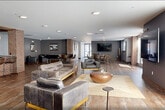 Located at 300 Riverfront Boulevard in Elmwood Park, NJ, this single-floor amenity space offers a generously sized communal environment designed for both relaxation and socializing. The expansive living room serves as the heart of the space, featuring warm hardwood-style flooring, leather seating arrangements, and large windows that flood the area with natural light and offer views of surrounding greenery. A striking exposed brick bar area with pendant lighting and bar seating adds an industrial-chic character, complementing the open-plan layout furnished with high-top tables and additional lounge seating. A dedicated game room provides entertainment with a pool table, foosball table, wall-mounted TV, and ample floor space, all finished with rich wood-tone flooring and neutral gray walls. Two well-appointed restrooms feature sleek stainless steel fixtures, stone-tile flooring, and clean, modern finishes. The outdoor patio extends the living space with a herringbone brick and stone-paved courtyard, outdoor dining furniture, and lush potted greenery, set against an attractive multi-story brick and siding building façade. This thoughtfully designed amenity space balances contemporary style with functional versatility.