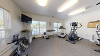 Located at 201 Millstone Rd in Florence, South Carolina, this single-floor fitness center offers a well-equipped workout space designed to support a range of exercise routines. The room features neutral white walls, carpet flooring, and bright overhead fluorescent lighting that creates a clean, functional atmosphere throughout.

Cardio equipment includes a treadmill, elliptical trainer, and a recumbent stationary bike, providing options for varied cardiovascular workouts. A multi-station cable and weight machine anchors the strength training area, complemented by a full dumbbell rack stocked with a wide range of free weights and additional loose weights and barbells for floor-based exercises. A wall-mounted flat-screen television is positioned for easy viewing during workouts, adding an entertainment element to the space.

Large windows along multiple walls allow generous natural light to fill the room while offering views of the surrounding outdoor area. A full-length wall mirror enhances the sense of openness and allows users to monitor their form during exercise. A glass-top utility stand provides a convenient surface for personal items. The space is accessible via a standard interior door marked with an exit sign, and a wall clock keeps users mindful of their time. Overall, this fitness center delivers a practical and comfortable environment for community members to maintain an active lifestyle.