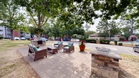 Located at 1521 E Franklin St in Chapel Hill, North Carolina, this outdoor community space serves as a welcoming gathering area within a residential complex. The space is anchored by a paved brick patio that provides a defined, ground-level area for relaxation and socializing. Comfortable wicker-style lounge chairs with teal cushions are arranged around a central stone fire pit or accent table, creating an inviting setting for residents to gather.

Mature, large-canopy trees shade the patio area, providing natural relief and a pleasant, park-like atmosphere throughout the warmer months. The lush, green tree cover contributes significantly to the overall ambiance, making the space feel tucked away and serene despite its proximity to parking areas and the surrounding multi-story residential buildings.

Large terracotta planters filled with vibrant tropical foliage and colorful flowering plants are positioned along the perimeter of the patio, adding decorative color and visual interest. The surrounding landscape is a mix of open grassy areas and tree-lined pavement, with the multi-story brick residential buildings of the complex visible in the background. The overall setting reflects a community-oriented outdoor environment that balances functional amenities with greenery and aesthetic detailing.