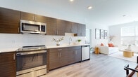 Located at 40 Bennett Rd in Englewood, NJ, this single-floor residence showcases a clean, contemporary design with an open layout and cohesive modern aesthetic. The kitchen serves as a central highlight, featuring dark wood-grain cabinetry extending from floor to ceiling, white quartz countertops, a classic subway tile backsplash, and a full suite of stainless steel appliances including a gas range, built-in microwave, dishwasher, and French door refrigerator. Light hardwood flooring flows seamlessly from the kitchen into the open living area, which is bathed in natural light from a large window overlooking greenery outside. The living space offers a bright, airy atmosphere with neutral white walls and recessed lighting overhead. Two well-proportioned bedrooms each feature large windows that invite abundant natural light and outdoor views. The primary bathroom includes a wide vanity with a white countertop, a large mirror, and a separate glass-enclosed shower. A second full bathroom mirrors the modern styling with tile flooring, a streamlined vanity with dark wood cabinetry, and a mirrored medicine cabinet. A dedicated closet with built-in shelving adds practical storage to the unit.