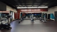 This single-level fitness center is designed to accommodate a comprehensive range of workout needs across multiple distinct zones. The cardio area is expansive, lined with a row of treadmills, elliptical trainers, stationary bikes, and rowing machines arranged along mirrored walls that amplify the sense of space. Overhead, exposed black-painted ceilings with industrial linear lighting create a high-energy atmosphere, while bold red accent panels add a striking visual identity to the space. The rubber flooring throughout provides durability and comfort underfoot across all workout areas. A dedicated strength training zone features a full suite of selectorized weight machines with red upholstery and white frames, along with free weight benches and cable systems. Wall-to-wall mirrors in this section allow users to monitor their form during exercises. A separate, more enclosed room offers additional strength equipment with neutral gray walls and softer recessed lighting, providing a quieter training environment. Towel stations and sanitation supplies are conveniently placed throughout the facility. The overall layout is thoughtfully organized to separate cardio and strength activities while maintaining an open, flowing floor plan that supports an efficient and motivating workout experience.