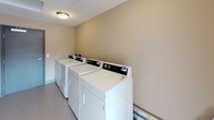 This shared laundry room is designed with practicality and functionality in mind. The space is organized into two distinct zones: a main laundry area and an adjoining hallway that extends toward a window at the far end. The laundry area houses a row of full-sized, commercial-grade top-loading washers and dryers equipped with digital control panels, lined neatly along one wall. The machines are finished in white and set against warm beige walls, creating a clean and orderly atmosphere. A metal-framed door with a hydraulic closer provides secure access to the main laundry section. Flush-mount ceiling light fixtures illuminate both areas evenly throughout. The hallway section features light-toned tile flooring that runs the length of the space, baseboard heating along one wall, and a built-in folding counter positioned near a window that allows natural light to filter in. A warm wood-toned door provides access to an adjoining room. The overall layout is straightforward and efficient, offering residents a comfortable, well-equipped environment for completing laundry with ease.