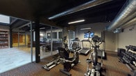 This resident amenities facility offers a well-rounded collection of spaces designed to support an active and social lifestyle. The central hub features a spacious entryway with large-format stone tile flooring, exposed structural beams, and soaring double-height ceilings that lend an open, airy feel to the space.

The fitness center is outfitted with a broad range of cardio equipment, including treadmills, ellipticals, a recumbent bike, and a rowing machine, along with a full dumbbell rack and weight bench. Exposed ductwork along the dark ceiling gives the gym an industrial aesthetic, while glass walls and doors maintain visual connectivity to adjacent areas.

The private screening room is a standout feature, furnished with rows of leather mission-style recliners facing a wall-mounted flat-screen display. Framed movie posters line the walls, enhancing the cinematic atmosphere.

The game room provides ample entertainment with a pool table, foosball table, and air hockey table, all set within a carpeted space with recessed lighting and exposed structural columns.

A cozy lounge area rounds out the amenities, furnished with rich leather sofas and armchairs, an area rug, and a built-in bookcase, creating a welcoming environment for reading or socializing. Together, these spaces form a versatile and well-appointed amenity suite.