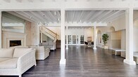Located along Pacific Coast Highway in Torrance, CA, this impressive two-story facility spans nearly 5,000 square feet across two floors, offering a well-appointed and versatile community space with a bright, coastal aesthetic. The ground floor opens into a sweeping, light-filled great room anchored by white-painted exposed beam ceilings, dark hardwood flooring, and crisp structural columns throughout. Wicker seating arrangements create comfortable lounge areas, while a grand piano takes center stage near floor-to-ceiling French doors and plantation-shuttered windows that invite abundant natural light. A library nook with built-in shelving, arched architectural details, and a reading table adds a refined touch. A marble-surround fireplace with mirrored accents and sputnik chandeliers enhances the upper lounge area, accessible by an elegant staircase. The second floor is notably more expansive and houses a dedicated fitness center equipped with cardio machines, weight benches, and full-length mirrors beneath a coffered ceiling with pendant fans. A billiards room features soaring ceilings, wall paneling, globe chandeliers, and two full-size pool tables. A bright multi-table gathering room with plantation shutters completes the upper level. Throughout, the space exudes a refined yet relaxed coastal character.
