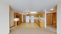 This single-floor space offers a functional and welcoming layout centered around a generously sized kitchen. The kitchen is outfitted with warm oak cabinetry that extends along multiple walls, providing an abundance of storage options. A full suite of white appliances—including a refrigerator, electric range, and built-in microwave—anchors the cooking area, while a double-basin sink is conveniently positioned along the countertop run. A dedicated built-in desk and cabinetry nook offers a practical workspace or organizational station just off the main kitchen area. Pendant and flush-mount lighting fixtures illuminate the space, contributing to a bright and functional atmosphere. The light-toned countertops complement the natural wood tones of the cabinetry throughout. The kitchen flows openly into an adjoining carpeted living area, creating a cohesive and connected layout that is well-suited for everyday living. The light-colored walls and ceiling enhance the sense of openness across the space. Overall, the design prioritizes practicality and comfort, with thoughtful storage solutions and a straightforward, clean aesthetic throughout.
