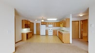 This single-floor space offers a functional and welcoming layout centered around a generously sized kitchen. The kitchen is outfitted with warm oak cabinetry that extends along multiple walls, providing an abundance of storage options. A full suite of white appliances—including a refrigerator, electric range, and built-in microwave—anchors the cooking area, while a double-basin sink is conveniently positioned along the countertop run. A dedicated built-in desk and cabinetry nook offers a practical workspace or organizational station just off the main kitchen area. Pendant and flush-mount lighting fixtures illuminate the space, contributing to a bright and functional atmosphere. The light-toned countertops complement the natural wood tones of the cabinetry throughout. The kitchen flows openly into an adjoining carpeted living area, creating a cohesive and connected layout that is well-suited for everyday living. The light-colored walls and ceiling enhance the sense of openness across the space. Overall, the design prioritizes practicality and comfort, with thoughtful storage solutions and a straightforward, clean aesthetic throughout.