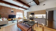 This single-floor loft-style space masterfully combines industrial character with contemporary finishes, creating a dynamic and highly functional living environment. The open-plan layout connects the living, dining, and kitchen areas seamlessly, anchored by soaring ceilings that showcase original dark wood beams and exposed ductwork running overhead.

The living area is defined by dramatic exposed brick walls, which extend to impressive heights and frame a large arched window that floods the space with natural light. Light hardwood flooring runs throughout, warming the industrial aesthetic. A ceiling fan complements the airy volume of the room.

The kitchen is outfitted with two-tone cabinetry—warm wood uppers paired with matte gray lowers—set against a marble-look backsplash and countertops. Stainless steel appliances, including a range, microwave, and refrigerator, complete the functional layout. The adjacent dining area accommodates a glass-top table with bench and chair seating.

The bedroom offers a calm, clean retreat with crisp white walls, recessed lighting, a ceiling fan, and light hardwood floors. A clerestory window adds subtle natural light.

The bathroom features floor-to-ceiling marble-look tile, a floating wood vanity, matte black fixtures, a glass-enclosed shower, and a backlit medicine cabinet, delivering a refined, spa-like finish.