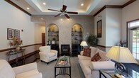 This single-floor clubhouse offers a sophisticated and inviting gathering space designed with comfort and elegance in mind. The entrance greets visitors with gleaming dark tile flooring, decorative columns, and large French doors with transom windows that flood the foyer with natural light and provide direct views of the outdoor pool area beyond.

The main lounge is a standout feature, showcasing a striking granite-clad accent wall flanked by two arched, built-in display niches with glass shelving. A central fireplace anchors the seating area, which is appointed with plush cream sofas and armchairs arranged around a glass-top coffee table. A coffered tray ceiling with a wood-blade ceiling fan adds architectural depth overhead, while rich wood crown molding runs throughout, reinforcing the refined aesthetic.

Adjacent to the lounge, a dining area features an elegant glass and wrought-iron table set beneath another tray ceiling with a ceiling fan, positioned near a kitchenette bar equipped with black appliances and rattan bar seating. A community information station with brochures and a bulletin board is conveniently accessible nearby.

Throughout the space, warm neutral tones, recessed lighting, decorative artwork, and lush indoor greenery contribute to a polished yet comfortable ambiance well-suited for community gatherings and social events.