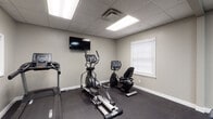 This single-level fitness facility is thoughtfully designed to accommodate a range of exercise needs in a clean, functional environment. The space features a dark rubberized floor that provides both durability and comfort underfoot, complemented by neutral gray walls and a drop-tile ceiling with recessed panel lighting that ensures bright, consistent illumination throughout.

The cardio area is outfitted with a treadmill, elliptical trainer, and a recumbent stationary bike, all positioned to allow easy access and movement. A wall-mounted flat-screen television is positioned above the cardio equipment, allowing users to stay entertained during workouts. A window with blinds admits natural light into the cardio section of the room.

On the opposite end of the space, a large floor-to-ceiling mirror expands the sense of depth and allows for form monitoring during exercise. This area is stocked with organized free-weight racks holding a full set of dumbbells, kettlebells, and resistance bands in various sizes, providing ample options for strength training. A seating area with blue chairs is available, suitable for group classes or rest between sets. A motivational wall decal reading "Start where you are. Use what you have. Do what you can." adds an encouraging atmosphere to the space.