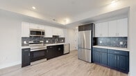 This thoughtfully designed single-floor space showcases a contemporary aesthetic throughout, with light hardwood-style flooring and a clean, neutral palette that creates a bright and cohesive atmosphere.

The kitchen is a standout centerpiece, featuring a striking two-tone cabinet design that pairs crisp white upper cabinets with dark lower cabinets, complemented by a separate section of slate-blue cabinetry. A bold black-and-white marble-look tile backsplash adds visual drama, while white quartz countertops provide a clean contrast. The kitchen is fully equipped with a complete suite of stainless steel appliances, including a range with microwave above, dishwasher, and a large French door refrigerator. Recessed lighting throughout ensures the space is well-lit.

The bathroom features a sleek, wall-mounted vanity in a natural wood finish with an integrated white sink and a mirrored medicine cabinet above. Rich, dark marble-look tile flooring adds a luxurious touch to the otherwise minimalist space.

A spacious room with corner windows allows abundant natural light and offers open views of the surrounding neighborhood streetscape, creating an airy and connected feel. The overall layout, as seen from the dollhouse view, flows logically between the open kitchen area and the additional rooms, making this a functional and visually appealing space.