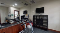 This fitness center is thoughtfully designed to support a comprehensive workout experience within a compact, single-level layout. The space is divided into distinct functional zones, each outfitted with purpose-specific equipment. The main area houses multiple cardio machines, including treadmills and stationary bikes, positioned alongside a large wall-mounted mirror that visually expands the room. A flat-screen television is mounted on the opposite wall, providing entertainment during workouts. Dark rubber flooring throughout the facility offers durability and safety underfoot.

A secondary area, separated by a low wood-paneled partition, features additional cardio and strength equipment such as a rowing machine, recumbent bike, and a weight bench. Large windows dressed with wood-framed blinds line multiple walls, allowing natural light to filter in while offering views of outdoor greenery. The wood trim detailing on the windows and baseboards adds a warm, traditional aesthetic to the otherwise functional space.

Near the entrance, a well-stocked dumbbell rack with color-coded free weights provides options for strength training. A wall-mounted hand sanitizer dispenser, informational signage, and a clock are also present near the glass-paneled entry door, contributing to the practical, user-focused design of the facility.
