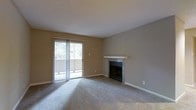 This single-floor residence presents a practical and comfortable layout with a cohesive neutral color palette throughout. The living area serves as the focal point of the space, highlighted by a brick-accented fireplace with a white mantel that adds a classic touch to the room. Plush wall-to-wall carpeting extends across the living area, contributing to a warm and inviting atmosphere. A sliding glass door fitted with vertical blinds opens to an outdoor balcony, allowing natural light to filter into the room while providing views of surrounding greenery.

Adjacent to the living area, a half-wall breakfast bar creates a natural transition into the kitchen zone, offering a casual dining or workspace surface. Built-in open shelving is integrated into the wall nearby, providing convenient storage and display options. Overhead HVAC vents are consistently placed throughout, ensuring climate control across the space. The textured ceilings and clean white baseboards complement the warm beige wall tones present in every room. The overall design is straightforward and functional, making efficient use of the available space with thoughtful built-in features.