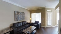 This single-floor, one-bedroom, one-bathroom unit delivers a thoughtfully designed living space with a clean, modern aesthetic. The living room serves as the centerpiece of the unit, featuring rich dark hardwood flooring that contrasts elegantly with the neutral taupe-toned walls. Crown molding adds a refined architectural detail, lending a polished finish to the space. A large frosted glass window allows natural light to filter into the room while maintaining privacy, creating a bright yet intimate atmosphere. The living area comfortably accommodates a full seating arrangement, as evidenced by the generous layout. A bold, mixed-media wall piece adds a distinctive decorative focal point, enhancing the room's visual character. The overall ambiance of the unit is warm and inviting, with a neutral color palette that provides a versatile backdrop for various décor styles. The single-floor layout ensures ease of movement throughout the space, making it a practical and comfortable option for everyday living.