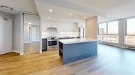 This well-appointed 2-bedroom, 1-bathroom unit offers a thoughtfully designed open-concept layout finished with light hardwood flooring throughout. The combined kitchen and living area forms the heart of the home, anchored by a striking blue kitchen island with a white quartz countertop and built-in shelving. The kitchen is equipped with white upper cabinetry, a subway tile backsplash, white quartz counters, a double-basin sink, and a full suite of stainless steel appliances including a French door refrigerator, range, and dishwasher. Floor-to-ceiling windows in the main living area flood the space with natural light while framing expansive outdoor views. A stacked in-unit washer and dryer is conveniently tucked within the living area. Both bedrooms are comfortably sized, each featuring large windows with outdoor views, light hardwood floors, and baseboard heating. The primary bedroom includes a sliding-door closet. The bathroom is finished with large-format marble-effect floor tiles, dark slate-tiled tub surround, a soaking tub, and clean white cabinetry. A hallway connects all rooms efficiently, completing this functional and modern layout.