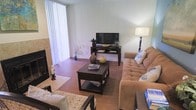 This two-story space offers a comfortable and well-appointed interior with a thoughtfully designed living area as its centerpiece. The living room features warm hardwood flooring that extends throughout, creating a cohesive and inviting atmosphere. A tiled fireplace anchors one side of the room, adding both a functional and decorative focal point. Large vertical-blind-covered windows allow generous natural light to filter in, brightening the space and creating an airy feel. The room comfortably accommodates a tufted upholstered sofa, accent chairs, and a dark wood coffee table styled with decorative accents, alongside a matching media console that houses the television. A tall floor lamp contributes to the warm ambient lighting. Neutral wall tones complement the earthy furnishings, giving the space a calm and balanced aesthetic. A patterned area rug adds visual texture underfoot. The overall design reflects a practical yet welcoming layout suited for everyday living and entertaining. With two floors, the space provides additional areas beyond the main living level, allowing for a clear functional separation between living and private spaces.