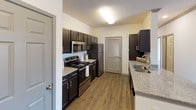 This single-floor, 2-bedroom, 2-bathroom residence offers a thoughtfully designed open-concept layout that maximizes both space and functionality. The living area features warm wood-look flooring, a ceiling fan, and direct access to a balcony through a large window that fills the room with natural light. A granite-topped kitchen island serves as the centerpiece connecting the living space to the kitchen, which is equipped with dark espresso cabinetry, granite countertops, stainless steel appliances including a refrigerator, microwave, and range, and a double-basin sink. The kitchen also includes a pantry with louvered doors for additional storage. Both bedrooms are carpeted and outfitted with ceiling fans for comfort. The primary bedroom features dual closets with built-in shelving, while the secondary bedroom includes a walk-in closet and a window with natural light. The two bathrooms are finished with matching dark cabinetry and granite vanity tops. The primary bath includes a standalone glass-enclosed shower, while the secondary bath offers a shower-tub combo. Throughout the residence, neutral tones and consistent finishes create a clean, cohesive aesthetic.