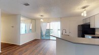 This single-floor residence offers a well-organized layout designed for comfortable everyday living. The open-concept living and dining area is anchored by rich hardwood-style flooring and bathed in natural light from a sliding glass door that leads to a private balcony, offering outdoor views. Sheer white curtains frame the windows and glass door, lending an airy, light-filled atmosphere to the space. A pendant light fixture adds a decorative touch above the dining area.

The kitchen features a central island with a built-in sink and light-toned countertops, complemented by white cabinetry and black appliances, including an electric range, microwave, and refrigerator. The space is both practical and efficient in its design.

The bedrooms are carpeted for comfort and include ceiling fans with light fixtures. The primary bedroom benefits from direct balcony access via a sliding glass door, as well as a walk-in closet with built-in shelving and an en-suite bathroom. The en-suite features a vanity with light countertops, blue cabinetry, and a shower-tub combination. A second full bathroom mirrors this clean, functional aesthetic with a tiled shower-tub combo and a well-lit vanity. Textured ceilings are present throughout, adding a consistent design detail across all rooms.