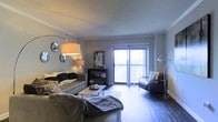 This single-floor space showcases a thoughtfully designed living area that blends comfort with contemporary style. The living room serves as the centerpiece, featuring rich hardwood flooring that extends throughout the space, contributing to a warm and cohesive aesthetic. Crown molding frames the textured ceiling, adding a refined architectural detail to the room.

Natural light floods the space through expansive sliding glass doors fitted with vertical blinds, which open to an outdoor balcony, offering both brightness and an indoor-outdoor connection. The room is generously sized, comfortably accommodating a full seating arrangement that includes a plush sofa, accent chairs, and a central coffee table.

Layered lighting enhances the ambiance, with a large arc floor lamp providing soft overhead illumination complemented by a secondary task lamp. A sleek console unit serves as a functional focal point along one wall, complete with decorative accessories and framed mirrors. The neutral grey and white color palette throughout creates a calm, modern atmosphere, while carefully chosen furnishings and accents add personality and depth to the space.