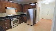 This single-floor space includes a well-appointed kitchen that serves as a central feature. The kitchen is outfitted with stainless steel appliances, including a gas range with oven and a full-size top-freezer refrigerator. Rich, dark wood cabinetry extends both above and below the countertops, providing generous storage space. The upper cabinets reach toward the ceiling, maximizing vertical storage capacity. Dark countertops contrast with the warm tones of the cabinetry, while light hardwood-style flooring runs throughout, lending a clean and cohesive look to the space. Recessed lighting in the kitchen ensures practical illumination for meal preparation. A hallway visible beyond the kitchen leads to additional rooms, suggesting a functional layout with clearly defined spaces. The light flooring and neutral wall tones contribute to an overall bright and airy ambiance. The kitchen's L-shaped configuration offers an efficient workflow, making good use of the available space while keeping a streamlined, practical aesthetic throughout.