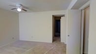 This single-floor space presents a straightforward and functional layout with a clean, neutral aesthetic throughout. The main room is finished with light beige walls and wall-to-wall carpeting in a muted tone, creating a understated, move-in-ready environment. A ceiling fan is mounted overhead, providing practical air circulation and modest overhead functionality. The room offers a generous sense of open floor space, well-suited for a variety of furniture arrangements.

An open doorway leads directly into an adjoining hallway or room, suggesting a connected interior layout that allows for easy flow between spaces. The transition between rooms is framed by a simple archway with clean-edged trim, consistent with the no-frills design approach seen throughout. The additional room visible through the doorway shares similar carpeting and wall finishes, maintaining a cohesive look across the space.

Electrical outlets are positioned along the baseboards, and a light switch is visible on the wall, reflecting standard residential utility placement. The overall ambiance is minimal and neutral, offering a blank canvas ready for personalization.