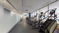 This fitness center is designed with both functionality and aesthetics in mind, offering a comprehensive workout environment across a single, elongated floor. The space is defined by a striking wall of floor-to-ceiling windows along one side, flooding the interior with natural light and providing open views of the surrounding exterior. The rubber-tiled flooring throughout provides a durable, slip-resistant surface ideal for high-traffic exercise use.

The cardio zone is prominently featured along the window-facing side of the room, lined with a row of treadmills, elliptical trainers, stair climbers, and stationary bikes — many equipped with individual digital displays. Opposite the cardio equipment, frosted glass partition walls lend the space a clean, modern aesthetic while subtly dividing areas without sacrificing openness.

Toward the rear of the facility, a dedicated strength training area houses a multi-station cable machine, a weight bench, a dumbbell rack with a full set of free weights, and additional resistance equipment. A wall-mounted mirror in this section enhances spatial perception and supports proper form during workouts. Wall-mounted flat-screen televisions are positioned throughout, providing entertainment and visual engagement during sessions.

The overall ambiance is clean, contemporary, and professional, with a neutral color palette of white, grey, and dark accents reinforced by recessed and fluorescent lighting.