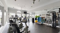 This fitness center offers a comprehensive workout environment within a single, efficiently laid-out floor. The space features classic architectural detailing, including crown molding, crisp white trim, and soft neutral-toned walls that create a clean, inviting atmosphere. Ceiling fans with recessed lighting provide comfortable air circulation and ample illumination throughout.

Large arched and rectangular windows line the walls, flooding the space with abundant natural light and offering views of the surrounding outdoor greenery. Full-length mirrors are strategically mounted along one wall, enhancing the sense of openness and allowing users to monitor their form during exercise.

The fitness floor is finished with durable rubber flooring suited for high-impact activity. Cardio equipment includes multiple treadmills, elliptical trainers, and both upright and recumbent stationary bikes. A multi-station cable strength machine anchors the free weights area, which is further stocked with a full rack of dumbbells and an assortment of medicine balls and stability balls. A weight bench rounds out the strength training offerings.

Interior glass windows and a connecting doorway provide a visual connection to an adjacent hallway, contributing to a sense of openness. The overall ambiance is bright, polished, and functional, reflecting a well-maintained facility designed to support a wide range of fitness activities.