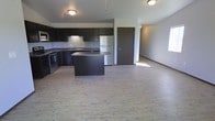 This single-floor space showcases an open-concept layout that seamlessly connects the kitchen and living area, creating a functional and cohesive environment. The kitchen is a standout feature, outfitted with dark espresso-toned cabinetry that extends to the upper and lower sections of the walls, providing generous storage. Stainless steel appliances, including a refrigerator and range, complement the contemporary aesthetic, while a central island with a dark countertop offers additional workspace and serves as a natural dividing point between the kitchen and the adjoining living area. The sink is positioned along the back counter, adding to the kitchen's practical layout. Light wood-style luxury vinyl flooring runs throughout the space, brightening the interior and creating a clean, modern feel that contrasts effectively with the darker cabinetry. Neutral gray walls contribute to the understated, versatile palette of the unit. Flush-mount ceiling light fixtures provide consistent illumination throughout. The open living area offers ample space for various furniture arrangements, and a doorway leads to an adjacent room, suggesting additional private spaces beyond the main living zone.
