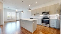 This well-appointed single-floor residence showcases a thoughtfully designed open-concept layout that seamlessly connects the kitchen and living areas. The kitchen is a standout feature, outfitted with grey shaker-style cabinetry, white quartz countertops, a classic subway tile backsplash, and a full suite of stainless steel appliances including a refrigerator, range, microwave, and dishwasher. A center island with pendant lighting serves as both a functional prep space and a natural gathering point. Warm hardwood-style flooring flows throughout the main living areas, complemented by recessed lighting and a ceiling fan. The living area is bathed in natural light through large windows and sliding glass doors that open to a private balcony. The bedroom offers plush carpeting, a bright window with outdoor views, and access to a bathroom featuring a glass-enclosed shower, a wide vanity with quartz countertops, and wood-look flooring. A generously sized walk-in closet with built-in shelving and hanging rods provides ample storage. The space combines clean, contemporary finishes with a functional floor plan, creating a bright and cohesive living environment throughout.