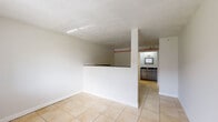 This single-floor unit offers a practical and efficient layout, encompassing a living room, two bathrooms, and a connecting hallway. Light beige ceramic tile flooring runs consistently throughout the entire space, contributing to a clean and cohesive aesthetic.

The living room is an open, simply finished area with white walls and a textured popcorn ceiling. A partial dividing wall with a dark countertop top separates the living area from the kitchenette, creating a semi-open floor plan. The entry area features a front door with a sidelight window that allows natural light to filter into the space.

The kitchenette is efficiently designed with white cabinetry, dark green granite countertops, a stainless steel sink, a built-in electric cooktop, an oven, and an over-the-range microwave. A framed pass-through window above the sink overlooks the living area, adding an open feel to the compact cooking space.

Both bathrooms are functional and neatly appointed. One bathroom features light gray large-format tile surrounding a standard bathtub and shower combination, a toilet, and a flush-mount ceiling light. Dark granite countertops are visible on cabinetry adjacent to the bathroom entry.

Overall, the space maintains a neutral, utilitarian character with consistent finishes, making it well-suited for practical everyday use.