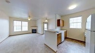 This single-floor space is designed with an open-concept layout that seamlessly connects the kitchen and living area, creating a functional and cohesive environment. The kitchen features a peninsula with a built-in sink and white countertops, providing both workspace and a natural divider between the cooking and living zones. Medium-toned wood cabinetry lines the upper and lower sections of the kitchen, offering ample storage, while a dishwasher is integrated beneath the counter. Tile flooring in the kitchen transitions to plush carpeting in the living area, distinguishing the two zones while maintaining an open feel. A pendant light hangs above the kitchen area, adding focused illumination. The living area is anchored by a fireplace with a traditional surround, serving as a focal point. A ceiling fan with a light fixture is centrally positioned, providing both air circulation and overhead lighting. Multiple windows on opposing walls allow natural light to fill the space from several angles. A doorway visible in the background suggests access to additional rooms or an exterior entry, adding to the overall flow of the layout.