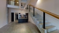 This two-floor space showcases a bold industrial aesthetic carried consistently throughout its design. The ground level features polished concrete flooring that extends seamlessly across the open layout, reflecting light and contributing to the spacious, loft-like atmosphere. Exposed ceiling infrastructure, including ductwork and pendant lighting, reinforces the industrial character while adding visual depth overhead.

A sleek kitchen anchors the main floor, outfitted with dark cabinetry, an island with an integrated sink, and under-cabinet lighting that provides a sharp contrast against the raw, utilitarian surroundings. The dark finishes of the kitchen create a dramatic focal point within the open space.

A prominent concrete staircase rises to the second floor, featuring black steel posts, clear glass panels, and a warm wood handrail — a combination that bridges industrial and contemporary design sensibilities. The upper level is partially visible from below, offering an open, connected feel between floors.

Natural and artificial light work together to brighten the interior, with recessed and pendant fixtures illuminating key areas. The overall ambiance is modern and minimalist, with a raw, edgy character defined by its material palette and open layout.