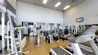 This single-floor fitness center offers a thoughtfully arranged workout space designed to accommodate a variety of exercise routines. The room features soaring ceilings that create an open, airy atmosphere, while a ceiling fan provides additional ventilation during intense workouts. Large mirrored panels line the walls, visually expanding the space and allowing users to monitor their form while exercising. The flooring consists of a durable, cushioned surface in a warm tan tone, providing both comfort and safety during workouts. The cardio section is well-stocked with elliptical machines, stationary bikes, and treadmills, offering ample options for aerobic training. Strength training is supported by a selection of cable and weight machines, along with a dedicated dumbbell rack positioned along the mirrored wall. Overhead, modern rectangular light fixtures ensure bright, even illumination throughout the space. The overall ambiance is clean, functional, and energizing, with a neutral color palette of gray and white that keeps the environment focused and distraction-free. The layout allows for easy movement between equipment stations.