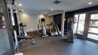 This fitness center offers a practical and functional space dedicated to health and wellness. The room is finished with durable dark rubber flooring, ideal for supporting heavy equipment and high-impact activity. Recessed lighting throughout the ceiling provides consistent, even illumination across the entire workout area.

The space is outfitted with a selection of cardio equipment, including treadmills, an elliptical trainer, and an upright stationary bike, giving users a variety of aerobic exercise options. A weight bench is also present for strength training. Large mirrors line one wall, enhancing the sense of space while allowing users to monitor their form during workouts.

The walls feature a two-tone design, with a deep navy accent wall providing a bold contrast against the lighter neutral walls. A window along one side of the room introduces natural light and contributes to an open, airy atmosphere. Ventilation is supported by ceiling vents, ensuring comfortable air circulation during exercise. The overall layout is thoughtfully arranged to allow users to move freely between equipment, making this fitness center a convenient and well-appointed facility.