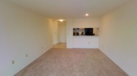 This single-floor residential unit offers a straightforward and functional layout designed for comfortable everyday living. The main living area is finished with neutral-toned wall-to-wall carpeting, creating a warm and inviting atmosphere. The space flows openly, providing ample room for a variety of furniture arrangements to suit different lifestyles.

A defining feature of this unit is the pass-through kitchen counter, which connects the kitchen to the living area. This design element promotes an open feel while maintaining a practical separation between the cooking and living zones. The kitchen is equipped with cabinetry in a natural wood finish and includes black appliances, giving it a classic and utilitarian character.

Adjacent to the living area, a tiled entryway or hallway leads to additional areas of the unit, with a door visible that likely connects to a closet or utility space. The transition from carpet to tile in this zone adds a natural delineation between spaces.

The interior is finished with clean white walls and ceilings, accented by recessed and flush-mount lighting that provides bright, even illumination throughout. The overall ambiance is clean, neutral, and move-in ready, making it a practical and versatile living space.