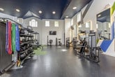 This single-floor fitness center is a comprehensively designed workout facility that seamlessly blends architectural character with professional-grade equipment. The space is defined by its dramatic vaulted ceilings finished in deep navy blue, contrasted against crisp white walls adorned with bold geometric graphic art in navy, blue, and green — adding an energetic, motivating atmosphere throughout.

The facility is thoughtfully divided into distinct functional zones. A spacious open floor area features green turf-style flooring ideal for functional training, complete with a multi-functional cable rig, battle ropes, and a built-in storage unit with cubbies. Adjacent to this, a dedicated strength training zone is outfitted with dark rubber flooring, a full rack of color-coded dumbbells, resistance bands, kettlebells, medicine balls, and multiple selectorized strength machines.

A cardio section houses treadmills, ellipticals, upright and spin bikes, and step platforms, all positioned beneath large arched and clerestory windows that invite generous natural light. Wall-mounted televisions are strategically placed throughout for entertainment. Large mirrors expand the visual depth of the space, while recessed lighting ensures bright, even illumination. The overall ambiance is polished, professional, and motivating.