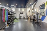 This single-floor fitness center is a comprehensively designed workout facility that seamlessly blends architectural character with professional-grade equipment. The space is defined by its dramatic vaulted ceilings finished in deep navy blue, contrasted against crisp white walls adorned with bold geometric graphic art in navy, blue, and green — adding an energetic, motivating atmosphere throughout.

The facility is thoughtfully divided into distinct functional zones. A spacious open floor area features green turf-style flooring ideal for functional training, complete with a multi-functional cable rig, battle ropes, and a built-in storage unit with cubbies. Adjacent to this, a dedicated strength training zone is outfitted with dark rubber flooring, a full rack of color-coded dumbbells, resistance bands, kettlebells, medicine balls, and multiple selectorized strength machines.

A cardio section houses treadmills, ellipticals, upright and spin bikes, and step platforms, all positioned beneath large arched and clerestory windows that invite generous natural light. Wall-mounted televisions are strategically placed throughout for entertainment. Large mirrors expand the visual depth of the space, while recessed lighting ensures bright, even illumination. The overall ambiance is polished, professional, and motivating.