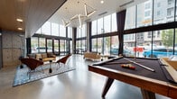 Dixon Place presents a striking first impression through its expansive lobby, defined by soaring double-height ceilings and an extensive curtain wall of floor-to-ceiling windows that flood the interior with natural light and offer sweeping views of the surrounding urban streetscape. Geometric, gold-toned triangular pendant lights descend dramatically from above, serving as a bold architectural focal point throughout the main lounge area.

The polished concrete floors extend throughout the space, lending an industrial-modern aesthetic that is balanced by warm furnishings, including tan and cognac leather sofas and sculptural accent chairs arranged atop patterned area rugs. A full-size pool table anchors the central gathering area, providing a social hub for residents.

A standout feature is a towering living moss wall installation adjacent to the Dixon Place signage, introducing a vibrant, organic element that contrasts richly against the dark stone-clad accent walls adorned with curated black-and-white historical photography. The reception area incorporates a warm wood-paneled desk set against exposed concrete columns, complementing the building's industrial roots. The overall ambiance is contemporary and community-focused, blending urban sophistication with inviting, lounge-style comfort.