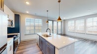 This single-floor space showcases a thoughtfully designed open layout with contemporary finishes throughout. A wide hallway with wood-look flooring and a paneled door leads into the heart of the home. The kitchen is a standout feature, centered around a generous island with a white quartz countertop, an undermount sink with a matte black faucet, and blue-gray cabinetry accented by gold hardware. White upper cabinets, a dark subway tile backsplash, and stainless steel appliances complete the culinary space. Dual gold pendant lights hang above the island, while multiple large windows with white blinds flood the area with natural light and offer outdoor views. The adjoining living area continues the wood-look flooring and provides a bright, open atmosphere. Bedrooms feature plush carpeting, ceiling fans, and well-sized windows. The bathrooms are elegantly finished with blue vanities, white quartz countertops, hexagonal stone-look tile flooring, and matte black fixtures. One bathroom includes a walk-in glass shower with white vertical tile, while another connects to a spacious walk-in closet. Recessed lighting and consistent neutral tones create a cohesive, modern ambiance throughout.