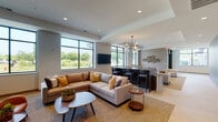 This single-floor space presents a thoughtfully designed amenity and common area layout with a contemporary aesthetic throughout. The main entrance features a striking glass-and-steel vestibule framed by floor-to-ceiling black-trimmed windows, flooding the entry with natural light and offering views of surrounding greenery. Large-format tile flooring in a warm, wood-tone finish extends through the corridors and entry areas, lending a cohesive, polished look.

A standout architectural feature is a substantial accent wall clad in textured, striated grey tile, creating a bold visual centerpiece that anchors the main circulation corridor. The hallways are generous in width, with recessed tray ceilings and recessed lighting adding depth and refinement.

The lounge area is a well-appointed social hub, furnished with a large sectional sofa, additional seating arrangements, and round coffee tables. A wall-mounted television and large windows overlooking lush outdoor scenery enhance the inviting atmosphere. Adjacent to the lounge is a bar counter with pendant chandelier lighting and barstool seating, ideal for gatherings.

Additional functional spaces include a dedicated package receiving room equipped with open metal shelving units and a commercial refrigerator for perishable deliveries. Utility corridors with grey steel doors provide organized access to service areas throughout the building.