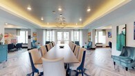 This thoughtfully designed single-floor amenity space offers a sophisticated blend of function and contemporary style. The centerpiece is a generously proportioned conference and dining room anchored by a long wood-topped table surrounded by upholstered seating. A striking sculptural gold chandelier hangs from a tray ceiling with warm ambient lighting, while floor-to-ceiling sheer curtains frame large windows that invite abundant natural light and views of the surrounding exterior. Herringbone-patterned wood-look flooring flows throughout the main areas, tying the spaces together with an elegant, modern aesthetic.

Adjacent to the main dining and conference area, an open lounge features plush teal velvet accent chairs, vibrant abstract artwork, and patterned area rugs, creating an inviting setting for casual gatherings. A kitchenette area offers a long white countertop island with industrial-style metal bar stools, pendant lighting, a shimmering tile backsplash, and coffee service, making it well-suited for social events or everyday use.

A dedicated media corner with wall-mounted television and a textured accent wall panel provides a relaxed seating nook. The space also includes two private meeting rooms finished with diamond-patterned wallcovering, carpeted floors, curated artwork, and flexible seating arrangements, ideal for small group gatherings or private sessions. Throughout, a cohesive palette of blues, golds, and neutrals lends the entire space a polished, resort-inspired character.