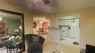 This single-floor residence showcases an open-concept layout that seamlessly integrates the living, dining, and kitchen areas into one cohesive space. The living area is anchored by a plush, dark upholstered accent chair paired with a decorative pillow, positioned beneath a ceiling fan that adds both function and character. Warm, neutral-toned walls and carpeted flooring create an inviting ambiance throughout.

The adjacent dining area features an elegant wrought-iron dining set beneath a chandelier, with a bold red artwork serving as a striking focal point on the wall. The space transitions naturally into the kitchen, which is defined by a peninsula counter that provides both prep space and a visual boundary between rooms. The kitchen is equipped with white appliances, including a range, built-in microwave, and refrigerator, complemented by light-toned cabinetry and tile flooring. A small decorative rug adds warmth to the kitchen floor. The layout promotes an easy flow between spaces, making the home well-suited for both everyday living and entertaining. Overall, the space offers a comfortable, functional design with consistent warm tones throughout.