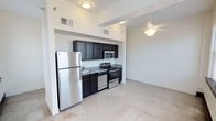 This single-floor unit offers a practical and well-organized layout designed for comfortable everyday living. The kitchen is a standout feature, equipped with a full suite of stainless steel appliances including a refrigerator, electric range, built-in microwave, and dishwasher. Rich, dark cabinetry extends from floor to ceiling, providing generous storage, while granite-style countertops add a polished finish. The kitchen flows openly into an adjacent living area, which features a ceiling fan overhead and concrete-style flooring that lends an urban, modern character to the space. Multiple rooms throughout the unit are bathed in natural light, thanks to large windows dressed with horizontal blinds and framed by dark wood trim that creates a striking visual contrast against the bright white walls. Each room is fitted with its own ceiling fan, supporting year-round comfort. The bathroom is clean and efficiently appointed, featuring a full tub-shower combination with chrome fixtures, a toilet, and a granite-accented vanity. Throughout the unit, high ceilings and an abundance of natural light from multiple window exposures contribute to an airy, open ambiance. The overall design balances functional simplicity with thoughtful detail.
