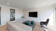 This thoughtfully designed single-floor space showcases a clean, minimalist aesthetic throughout its open-plan layout. Light hardwood flooring runs seamlessly across the entire space, contributing to a warm yet modern atmosphere.

The living area is anchored by a large, modular sectional sofa in neutral tones, paired with teal accent ottomans that introduce a subtle pop of color. Recessed lighting illuminates the space, while a wall-mounted flat-screen television serves as a focal point. Dark-framed windows allow natural light to filter in, offering views of the surrounding urban environment.

The kitchen is a standout feature, designed with both functionality and style in mind. Crisp white cabinetry lines the walls, complemented by a textured concrete-look backsplash that adds depth and character. A generous island with a pristine white countertop provides ample prep space and casual seating for several guests, with a sleek linear pendant light suspended above. Professional-grade appliances, including a range with a stainless steel hood, complete this well-appointed culinary space.

A dining area with a long communal table bridges the kitchen and living spaces, ideal for gathering and entertaining. Large glass doors lead to an outdoor terrace, further enhancing the sense of openness and connection to the outdoors. Overall, the space exudes a refined, contemporary sensibility.