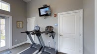 Located at 6300 Caleb's Creek Way in Westerville, Ohio, this fitness center offers a clean and functional workout environment designed to support an active lifestyle. The space is finished with neutral gray walls and carpeted flooring, creating a practical yet comfortable atmosphere for exercise. The cardio area is equipped with multiple treadmills, each featuring individual digital display consoles for tracking workout metrics. A wall-mounted flat-screen television is positioned above the equipment, allowing residents or members to stay entertained during their workout sessions. The room is well-organized, with clearly marked accessible restroom facilities conveniently located directly within the fitness space, ensuring ease of access for all users. Accessibility signage is prominently displayed throughout, reflecting a commitment to inclusive design. The single-floor layout provides a straightforward, no-frills environment focused on delivering essential fitness amenities. The overall ambiance is tidy and purposeful, making it a practical resource for daily exercise routines.