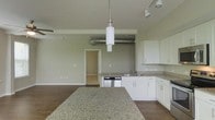 This single-floor space showcases a thoughtfully designed open-concept layout that effortlessly blends modern and industrial aesthetics. The kitchen serves as the heart of the space, featuring crisp white cabinetry, granite countertops, and a stainless steel dishwasher. A large granite-topped island anchors the kitchen area, offering ample workspace and serving as a natural divider between the cooking and living zones. Pendant lighting hangs above, complementing the track lighting overhead to provide well-distributed illumination throughout. A defining industrial element is the exposed metal ductwork that runs across the ceiling, adding character and a loft-like ambiance to the space. The open living area extends seamlessly from the kitchen, finished with rich dark hardwood flooring that contrasts beautifully with the light gray walls and white trim. The generous ceiling height enhances the overall sense of openness and airiness. A doorway visible in the background hints at additional rooms, suggesting a functional and well-connected floor plan. The overall aesthetic is clean, contemporary, and versatile, making this space well-suited for a variety of lifestyles.