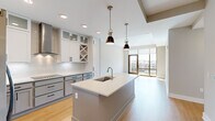 Located at 350 Carraway Crossing in Chapel Hill, NC, this thoughtfully designed one-bedroom, three-bathroom residence offers a contemporary single-floor layout that maximizes both function and style. The open-concept kitchen is a true centerpiece, showcasing white shaker cabinetry with glass-front uppers, a built-in wine rack, quartz countertops, a subway tile backsplash, stainless steel appliances including a wall oven and microwave, an induction cooktop, and a large center island with a undermount sink — all illuminated by stylish pendant lighting. The living room flows seamlessly from the kitchen and is bathed in natural light through expansive floor-to-ceiling windows and a glass door that opens to an outdoor balcony with tree-lined views. The bedroom is generously proportioned, featuring plush carpet, large windows with outdoor views, and direct access to a well-appointed en suite bathroom complete with a dual-sink vanity, dark hardware accents, and an adjacent walk-in closet with multi-level shelving and hanging rods. An in-unit laundry area houses a full-size front-loading washer and dryer. Light hardwood flooring flows through the main living areas, tying the space together with a clean, modern aesthetic.