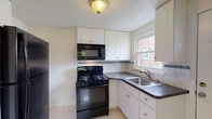 This two-floor home offers a well-organized layout with distinct living spaces across both levels. The main floor welcomes visitors through a front entry door into a bright living room adorned with warm hardwood flooring and crown molding trim, with a staircase leading to the upper level. Adjacent to the living area is a compact, efficiently designed kitchen featuring white shaker-style cabinetry, dark countertops, white subway tile backsplash with a decorative mosaic accent border, black appliances including a gas range and over-the-range microwave, and a stainless steel sink beneath a window that allows natural light to flow in. A separate room on the main floor features light tile flooring, crown molding, and a large window overlooking a green outdoor area. Upstairs, the layout — as seen in the dollhouse view — includes multiple rooms with gleaming hardwood floors throughout. The bedrooms are bright and simply finished with white walls and windows framing views of the surrounding brick neighborhood. The bathroom is a standout with its retro character, showcasing pink ceramic wall tiles accented by black border tiles, a full soaking tub with shower, and a pedestal-style sink with a wall mirror. Throughout the home, natural light is a consistent feature, contributing to an airy and inviting atmosphere.